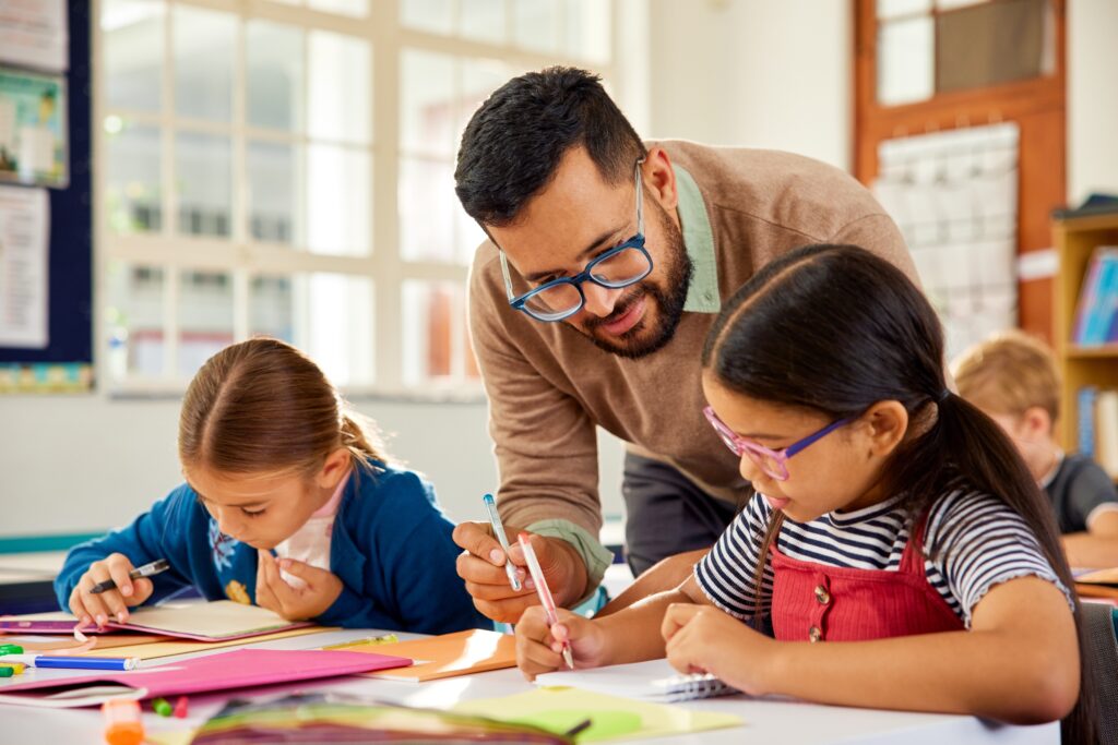 Male,Teacher,In,Classroom,Helping,School,Kids,At,Desk.,Little